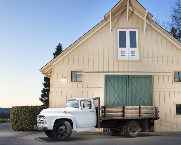 A vintage white truck is parked in front of a light-colored barn with green doors, under a clear sky.