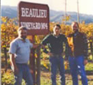 Three men stand in front of a sign that reads "Beaulieu Vineyard, Napa" with vineyards and mountains in the background.