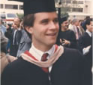 A young person wearing a graduation cap and gown stands outdoors, smiling amidst a crowd of people.