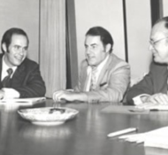 Three men in suits engage in a discussion around a large wooden table, with papers and a dish in the center.