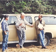 Three men stand and converse next to a tan SUV in an outdoor setting with greenery in the background.