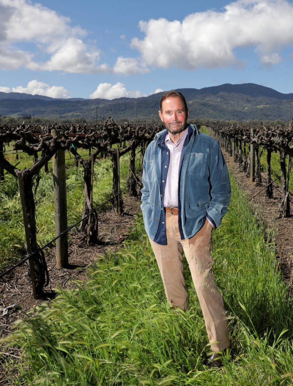 A man in a blue jacket and beige pants stands in a vineyard with mountains in the background under a partly cloudy sky.