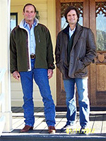 Two men standing in front of a wooden door on a porch, both wearing jackets and jeans.