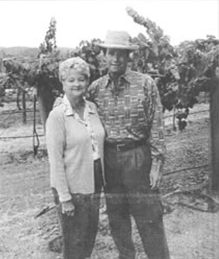 An elderly man and woman stand together outdoors in front of grapevines. The man wears a hat and patterned shirt, and the woman wears a light-colored cardigan over a shirt.